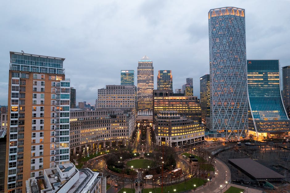 A wide-angle view of Canary Wharf's downtown skyline during early evening with illuminated office buildings, including the distinctive twisting cylindrical tower on the right. In the foreground, a residential building with multiple balconies and large windows is visible. Below, a small park with pathways, green grass, and street lamps is situated at the base of the high-rise structures. The scene shows the typical cityscape associated with professional real estate and corporate relocations, supporting the context of house and office removals. The lighting indicates dusk or early evening, and the overall scene reflects the urban environment where furniture transport and packing for home relocation services provided by Man with Van Canary Wharf are often conducted.