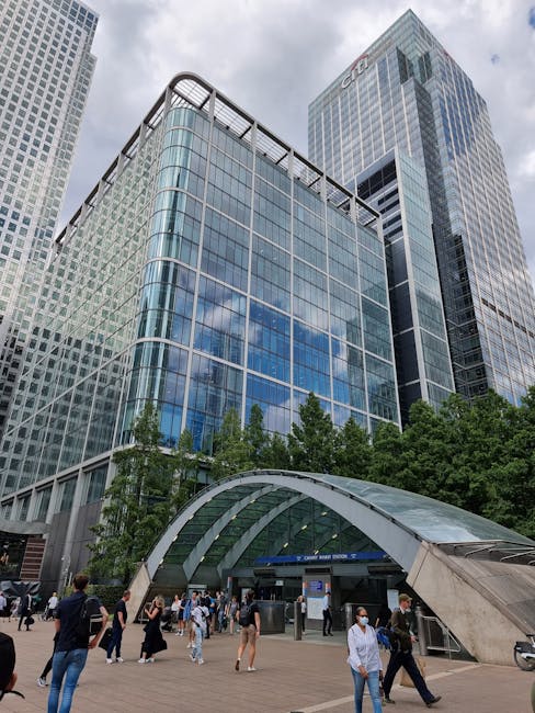 A modern, multi-storey office building with a glass facade reflecting the sky and surrounding structures, located in Canary Wharf. In the foreground, a group of people are walking on the paved plaza near the entrance, some wearing face masks. The entrance features a curved, transparent canopy made of glass and metal, leading into the building. Several trees and greenery are visible at the base of the building, adding natural elements to the urban environment. This setting depicts a busy commercial area suitable for house removals and furniture transport services as part of a home relocation or moving process, with the building's towering height and glass exterior providing a clear view of downtown Canary Wharf. Man with Van Canary Wharf frequently facilitates such moves, including loading and unloading furniture and boxes in this area.