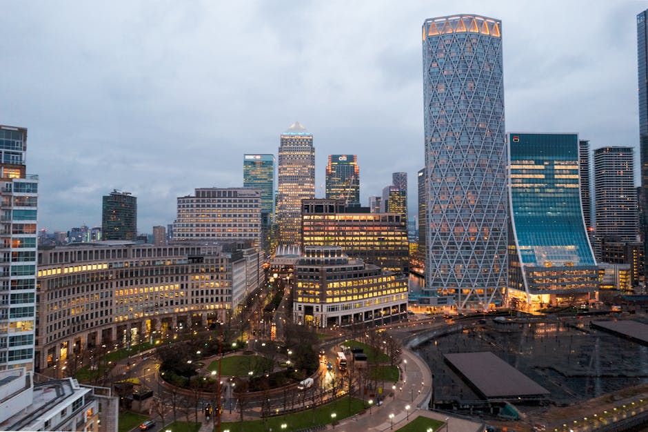 A wide-angle view of Canary Wharf's downtown skyline during early evening with illuminated office buildings, including the distinctive twisting cylindrical tower on the right. In the foreground, a residential building with multiple balconies and large windows is visible. Below, a small park with pathways, green grass, and street lamps is situated at the base of the high-rise structures. The scene shows the typical cityscape associated with professional real estate and corporate relocations, supporting the context of house and office removals. The lighting indicates dusk or early evening, and the overall scene reflects the urban environment where furniture transport and packing for home relocation services provided by Man with Van Canary Wharf are often conducted.