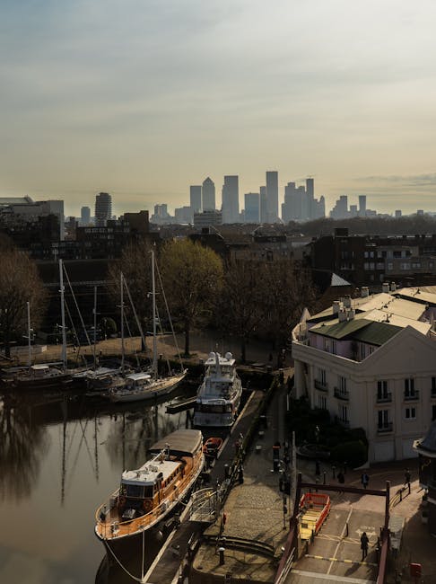 Aerial view of Canary Wharf in London during evening with modern high-rise office buildings and residential apartments lining the riverbank, with some windows illuminated and the sky overcast. In the 